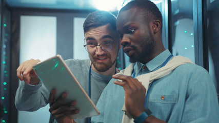 Close-up multi-race friendly engineers in data center using smartphone browsing social media showing funny grimaces coworking in server room.