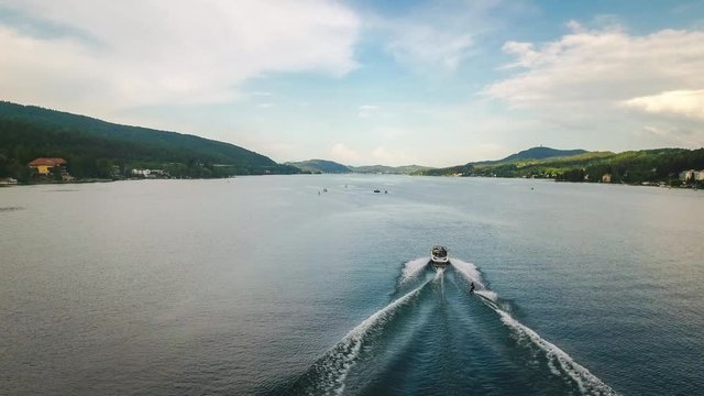 Aerial drone tracking shot of a speed boat and a man waterskiing doing water sports in Velden Am Worthersee lake, Austria on a beautiful sunny day mountain view 