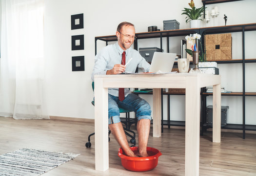  Comic modern office table situation. Businessman typing on laptop keyboard and soaring his feet in Foot hot Bath under table. Distance work in worldwide quarantine time concept image.