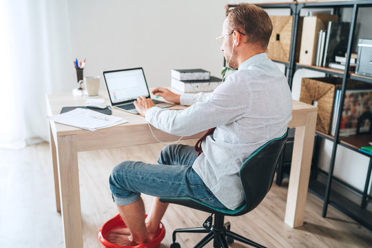  Comic modern office table situation. Businessman typing on laptop keyboard and soaring his feet in Foot hot Bath under table. Distance work in worldwide quarantine time concept image.