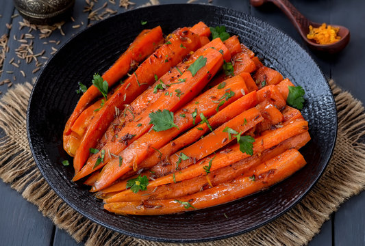 Braised carrots with cumin and orange in a black plate. Dark wooden background, selective focus.