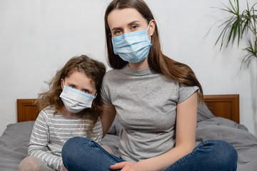 Close up front view young mother and little daughter wear facial mask, look at camera feels afraid, sitting on bed at home. COVID-19 sars-cov-2 epidemic pandemic virus outbreak protection concept