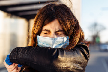 Young girl sneezes in a medical sterile mask on a bus station. She coughs into elbow. Public...