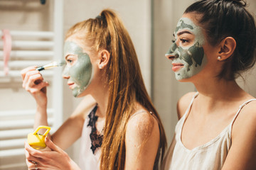 Caucasian woman with red hair and her brunette friend are applying a facial anti acne mask while...