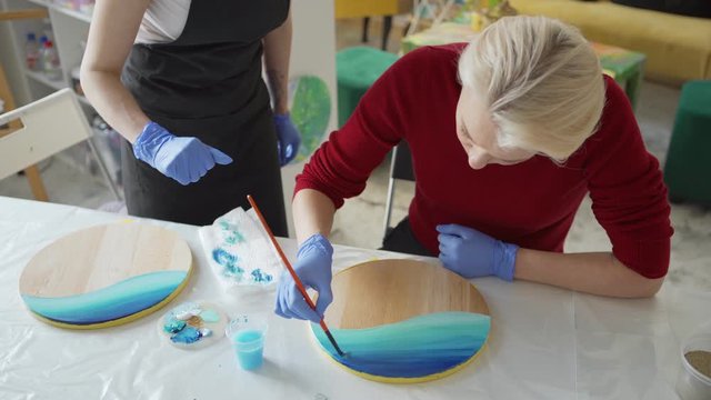 From above shot of art teacher explaining middle aged female student how to create sea painting with acrylic paints on wooden round canvas in art studio
