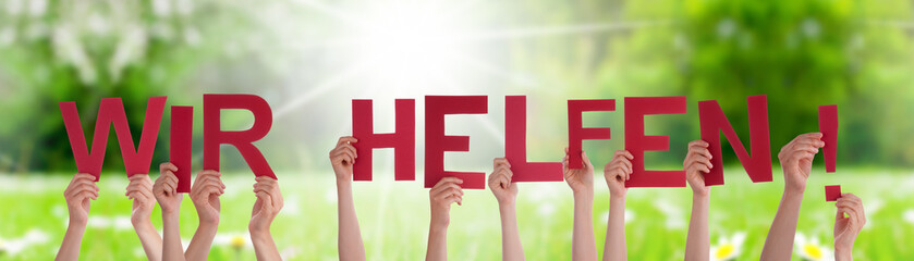 People Hands Holding Colorful German Word Wir Helfen Means We Help. Sunny Green Grass Meadow As...