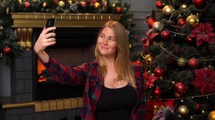 Young beautiful girl or woman in a red checkered shirt taking selfie with christmas tree on background.