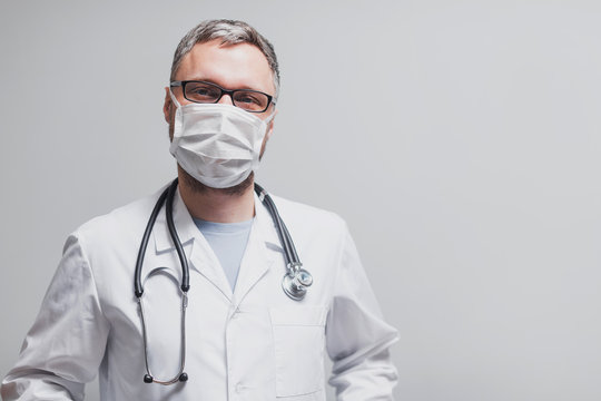 doctor in a lab coat stands in a medical mask on a light background on a bright day