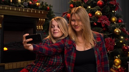 Two pretty young girls or women are sitting near a New Year or Christmas tree and taking selfie on a mobile phone.