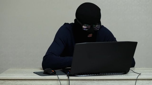 Funny worried male hacker in black mask balaclava puts on glasses and do hacker attack on laptop