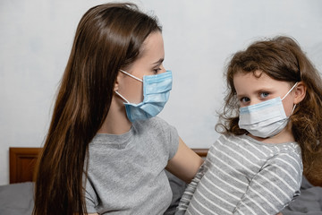 Loving pretty young  mother looking at little daughter wearing protective mask sitting on bed at home. Home quarantine. Concept of coronavirus or COVID-19 pandemic disease symptoms