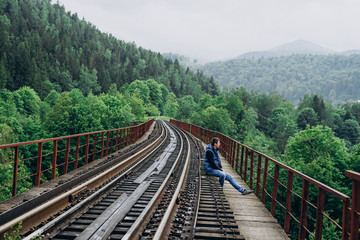 Fototapeta na wymiar A man sits on a railway track in a blue jacket. Track on mountains background.