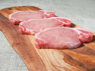 Three raw uncooked pork chops on a wooden cutting board,