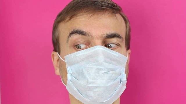 front view on male face in medical mask, looks around with fear and distrust then close eyes in panic, quarantine or epidemic of virus or protection of pollution, pink background