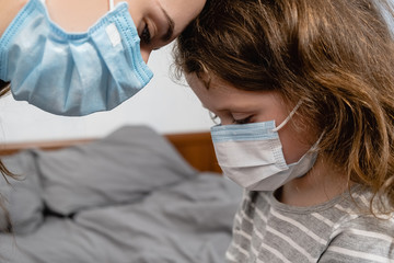 Close up of caucasian mother and little daughter wearing protective mask sitting on bed at home. Self isolation, home quarantine. Concept of coronavirus or COVID-19 pandemic disease symptoms