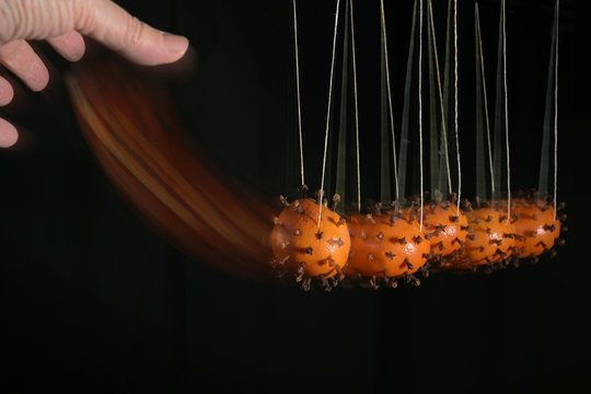 Hand is moving newton's cradle with coronavirus models from tangerines peppered with cloves, to illustrate how the infection is transmitted by contact, motion blur, black background, copy space