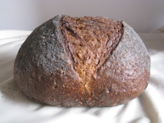 Pan de centeno obscuro cubierto con semillas de forma de bollo ovalado con grieta de hornear en centro sobre mantel blanco