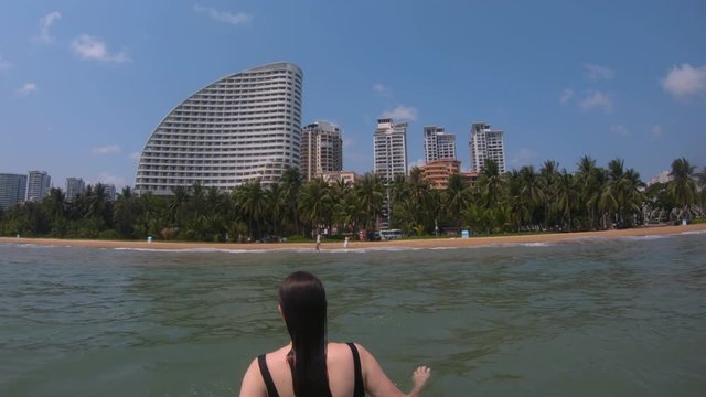 A young girl swims in the sea in China. A woman is standing waist-deep in blue water. Strong waves, tall palm trees, large buildings on the shore of Hainan Island.