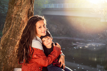 Beautiful mom with her son, sitting on a bench in the park, river behind them.