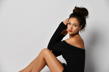 Fototapeta premium Portrait of gorgeous woman in stylish black tight dress sitting sideways to us with her legs tucked up and looking at us
