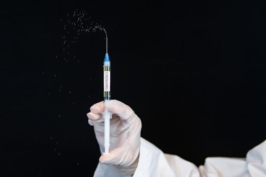 Selective focus closeup, syringe labeled coronavirus, squirting drops of liquid being held by a gloved hand vertically over a black background 