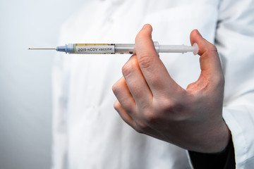 Selective focus closeup syringe filled with 2019-nCOV flu vaccine held by bare hand. Background of out of focus white lab coat. copy space bottom left