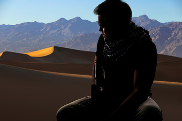 Silhouette of a male soldier resting in the shade on a desert and holding a rifle.  Depicts the...