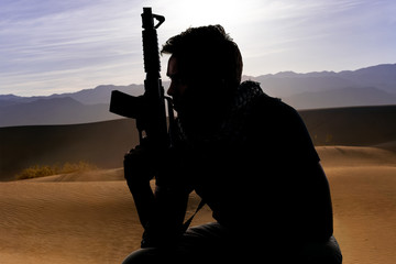 Silhouette of a male soldier resting in the shade on a desert and holding a rifle.  Depicts the...
