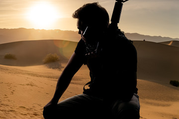 Silhouette of a male soldier resting in the shade on a desert and holding a rifle.  Depicts the...