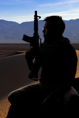 Silhouette of a male soldier resting in the shade on a desert and holding a rifle.  Depicts the...