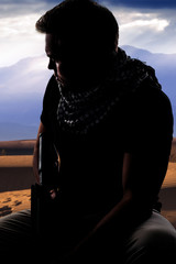 Silhouette of a male soldier resting in the shade on a desert and holding a rifle.  Depicts the...
