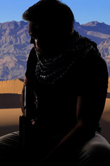 Silhouette of a male soldier resting in the shade on a desert and holding a rifle.  Depicts the...