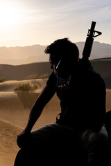 Silhouette of a male soldier resting in the shade on a desert and holding a rifle.  Depicts the...