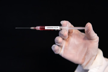 Selective focus closeup, syringe labeled coronavirus vaccine holding red liquid being held by a gloved hand horizontally over a black background 