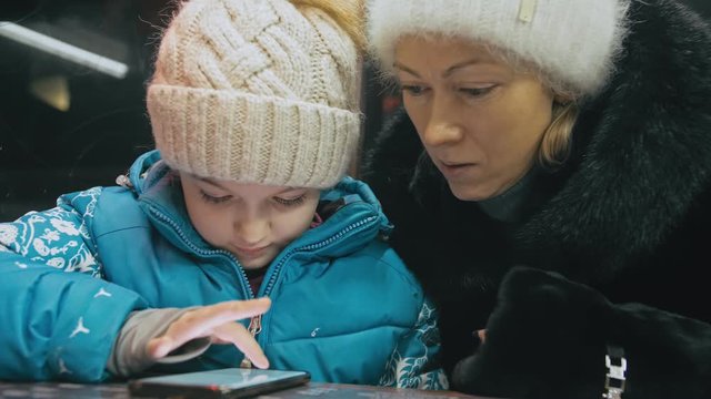 Attractive caucasian mother and daughter using smartphone sitting in winter cafe in jacket, hat. Advanced little child girl shows and explains to mom woman on smartphone, touching and watching.