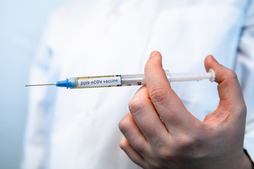 Selective focus closeup syringe filled with 2019-nCOV flu vaccine held by bare hand. Behind out of focus white lab coat over white back ground