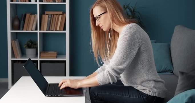 Side view of competent female worker in eyeglasses and casual outfit using laptop while sitting at couch. Mature woman with blond hair working at home.