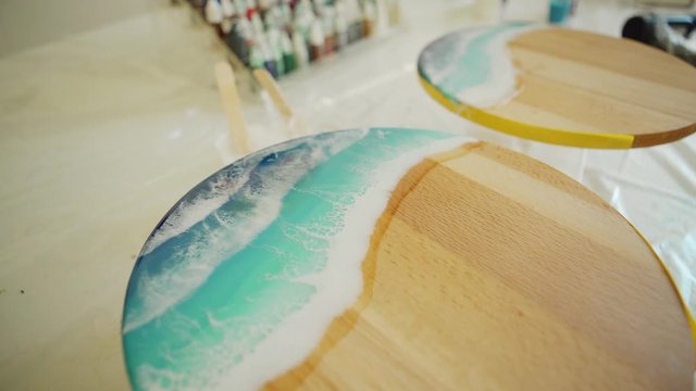 Tracking from above closeup shot of table with art equipment in art studio and two unfinished fluid sea paintings made with epoxy resin and acrylic paints on round wooden canvases