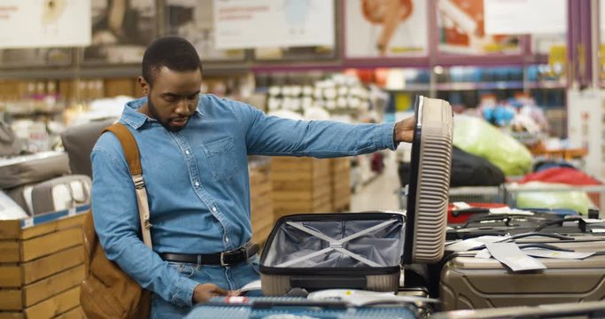 Handsome young African American man choosing suitcase with wheels in hypermarket. Good looking stylish cheerful guy trying cases in big shop. Male shopper in supermarket buying bag.