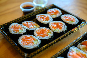 Salmon maki roll sushi on a black plastic tray, side with soy sauce and wasabi Is a set of dishes delivered on an orange wooden table.