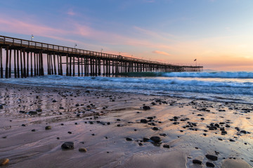 Obraz premium Ventura Pier at sunset, Ventura, California. Rocks and sand in foreground, water receded in low tide, with incoming waves. Lamps on pier; colored twilight sky in background.