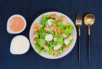 Bowl of healthy green vegetable salad on top with quail egg and biscuits. Side dish with spoon, fork, white cream and thousand island salad dressing