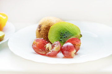 On the plate is a half of a very ripe juicy green kiwi with grapes on a white plate. Close up. Light monochrome background