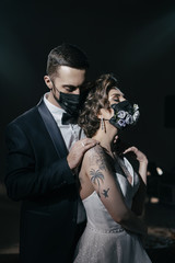 The bride and groom in wedding dresses and in black medical masks decorated with fresh flowers