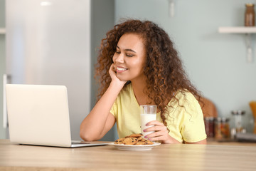 Young African-American woman with laptop drinking milk in kitchen