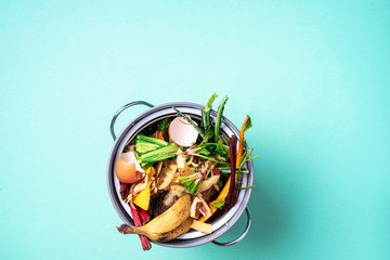 Peeled vegetables on chopping board, white compost bin on blue background. Top view of kitchen food...