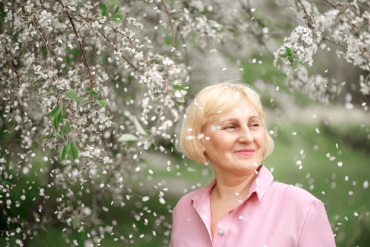 Happy Handsome Smiling Dreaming Older Woman With Short Blonde Hair In Pink Dress. Spring Park With Blooming Cherry. Falling Petals. Front View.