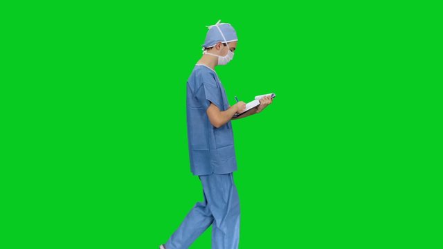 Side view of young doctor in mack and uniform writing on clipboard while walking against green screen background, Chroma key 