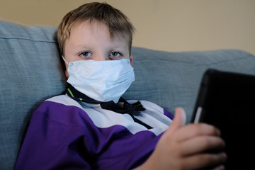 Schoolboy wearing a mask looks at the camera while holding a device at home during the Covid-19 / coronavirus pandemic