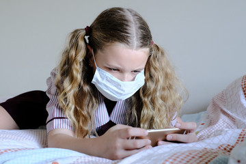 Schoolgirl wearing surgical mask does homework on a device during Covid-19 / coronavirus pandemic